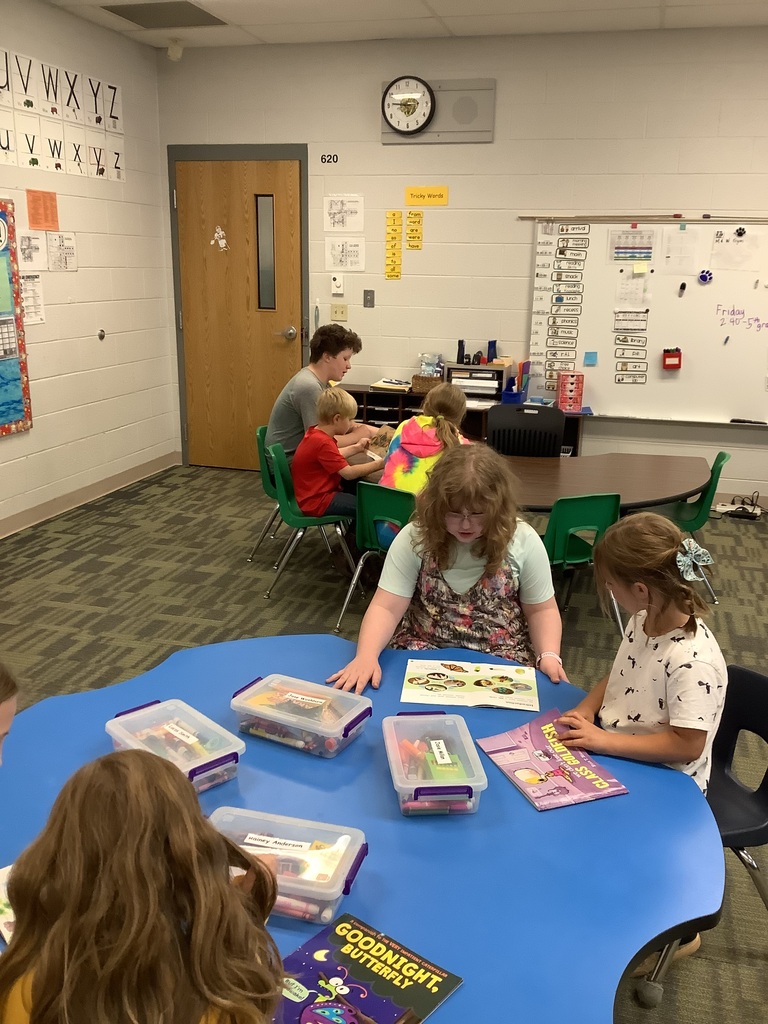 Students working in reading pairs at tables inside a classroom. In the foreground, an older girl and a younger girl sit together at a blue clover-shaped table, looking at open books alongside clear storage bins. In the background, other pairs of students sit around a brown kidney-shaped table with classroom whiteboards, alphabet strips, and schedules mounted on the wall.