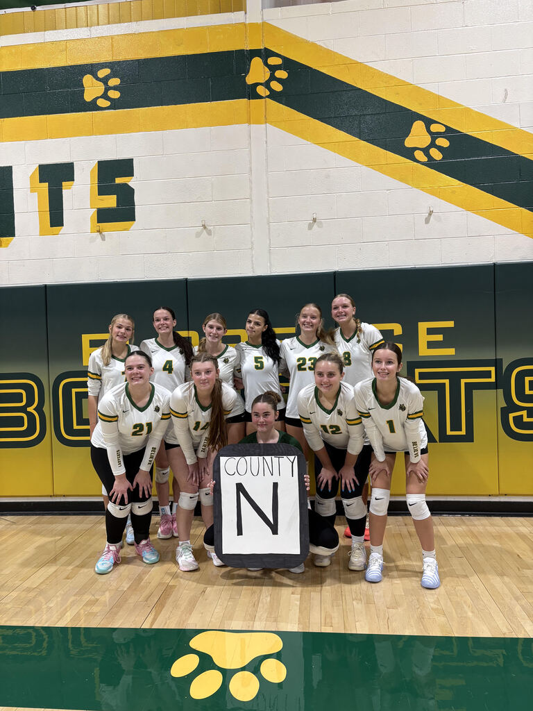 A high school girls' volleyball team posing for a team photo on a wooden basketball court inside a gymnasium, with green, gold, and white wall decorations behind them.