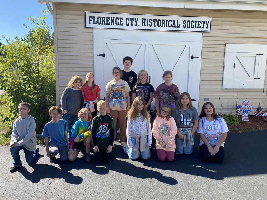 A group of 13 children pose together outdoors in front of a light tan building with a sign that reads "FLORENCE CTY. HISTORICAL SOCIETY". Behind them are large white wooden barn-style double doors. The children are arranged in two rows—some kneeling or sitting on the pavement in the front row, and others standing in the back row. They are wearing casual school clothes, such as t-shirts, hoodies, and jeans, enjoying a sunny day outdoors.