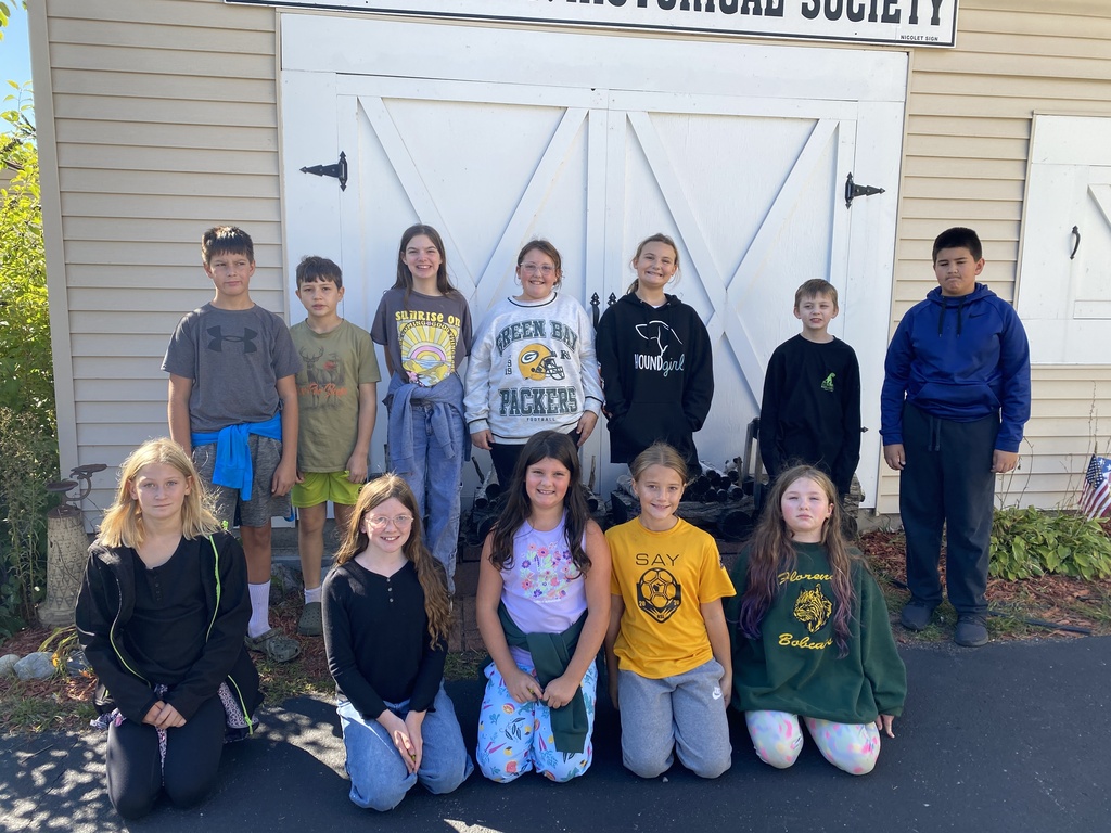 A group of 12 young students posing outdoors in front of a tan building with large white barn doors. Above the doors, a sign reads "...HISTORICAL SOCIETY". The children are arranged in two rows: seven standing in the back row and five kneeling in the front row. They are wearing casual clothing, including graphic t-shirts, sweatshirts (including a Green Bay Packers sweatshirt and a Florence Bobcats sweater), and jeans.
