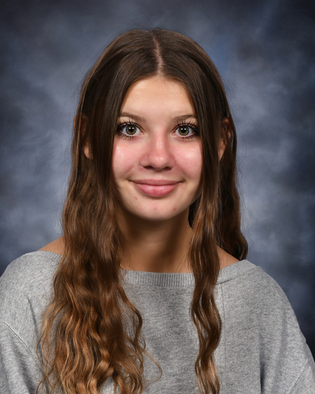 A headshot photograph of a young female with long, wavy light-brown hair styled with a center part. She is wearing a light grey off-the-shoulder sweater and smiling warmly into the camera against a textured blue studio backdrop.