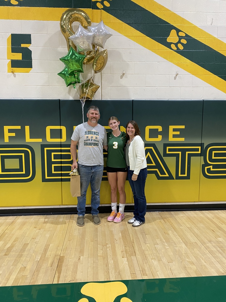 A young female volleyball player in a green jersey with the number 3 standing on a gymnasium floor with her parents. They are posing in front of a green and gold wall that reads "FLORENCE BOBCATS" while holding a bouquet of balloons celebrating senior night.