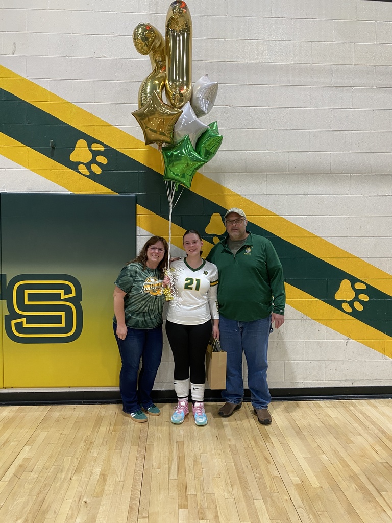 High school volleyball player Quinn Cassidy wearing a white number 21 jersey poses with her parents on a gymnasium basketball court during senior night, holding a bouquet of green, gold, and silver balloons including gold "21" number balloons.