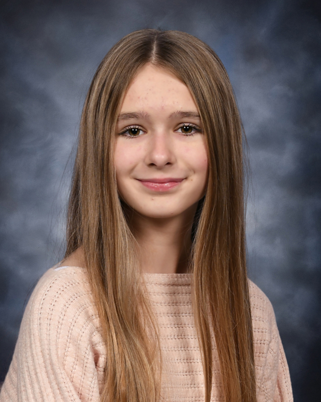 School portrait of a young girl with long, straight light brown hair, smiling gently in front of a mottled blue backdrop while wearing a light pink knit sweater.