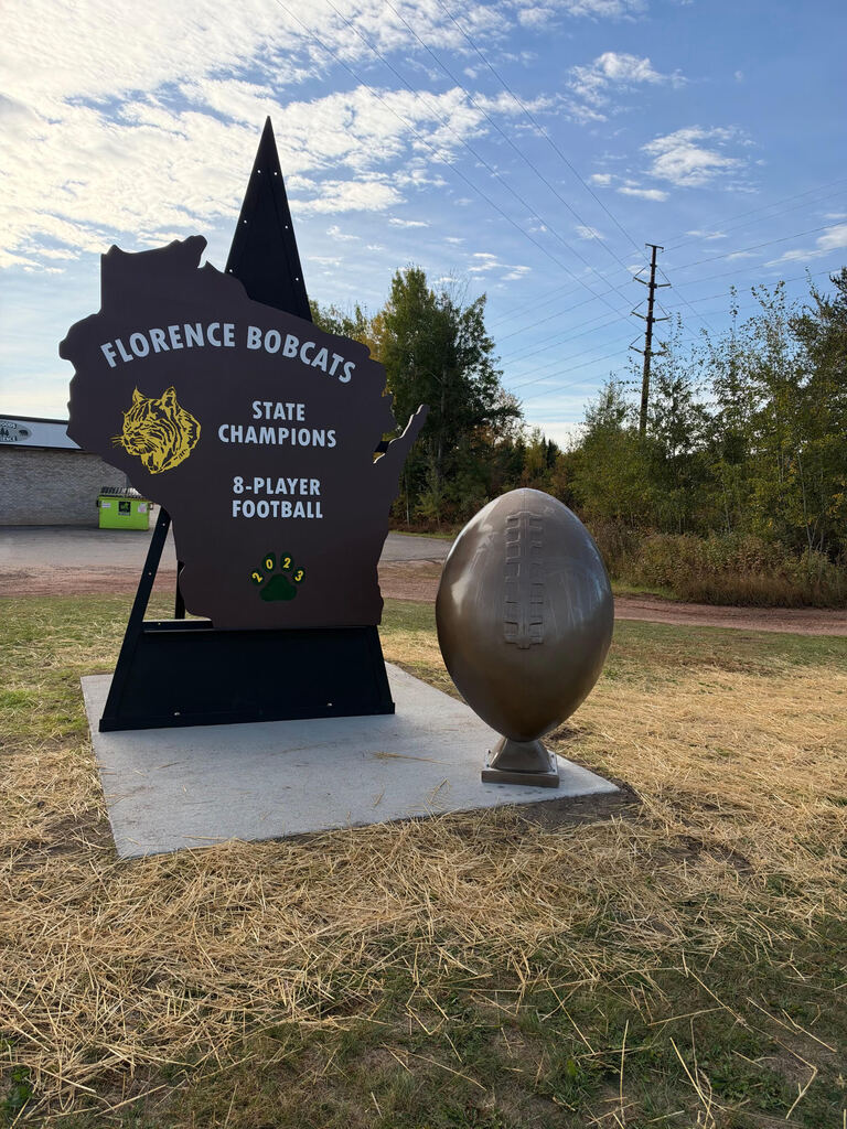 A brown monument shaped like the state of Wisconsin reading "Florence Bobcats State Champions 8-Player Football 2023" next to a large bronze football sculpture.