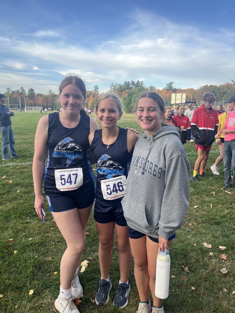 Three young female athletes smiling together outdoors on a grassy field during a cross-country meet under a partly cloudy sky. The two girls on the left wear matching navy blue "Northwoods Storm" sleeveless athletic uniforms with white race bibs numbered 547 and 546 attached to the front. The girl on the right wears a grey hooded sweatshirt reading "Indian Shores Florida" over dark shorts and holds a light-colored water bottle. Other runners and spectators are visible on the grass field in the background.   