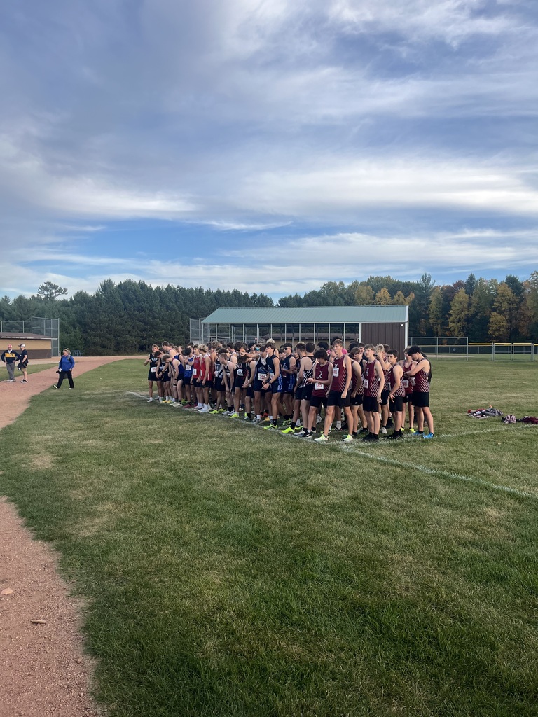 A large group of high school cross-country runners lined up along a white starting line on a grassy field, waiting for the start of a race. The runners wear various colorful team uniforms, including maroon, blue, and red singlets with bib numbers. In the background stands an athletic facility building, a pine forest, and autumn trees under a wide, wispy blue sky.   