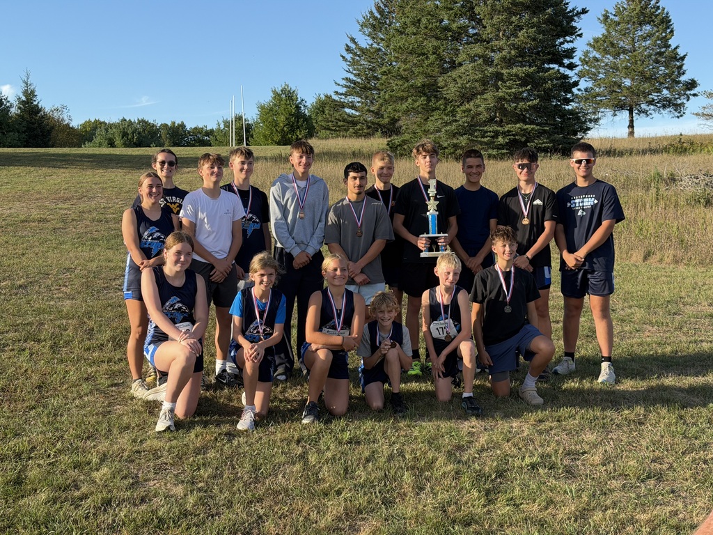 A group of young cross country team members pose outdoors on a grassy hill on a clear, sunny day. The back row features eleven team members standing, with one holding a tall first-place trophy and several wearing medals around their necks. The front row consists of six team members kneeling on the grass, wearing athletic gear and medals. Large pine trees and a blue sky form the background.   