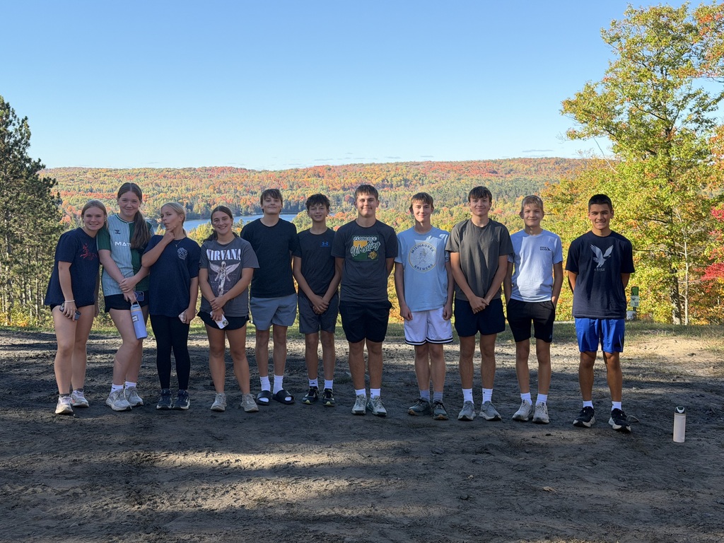 A group of ten young people pose standing side-by-side outdoors on a dirt path under a clear blue sky. Behind them, a scenic overlook reveals rolling hills covered in colorful autumn foliage and a lake in the distance. The group wears casual athletic t-shirts and shorts, with a water bottle resting on the ground to the right.   