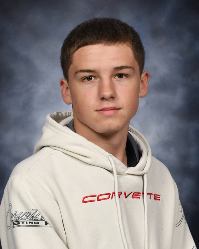 A portrait of a young man with short brown hair posing against a mottled gray studio backdrop. He is wearing an off-white hooded sweatshirt with the word "CORVETTE" printed in red across the chest, and a logo reading "Corvette Stingray" printed on the sleeve.   