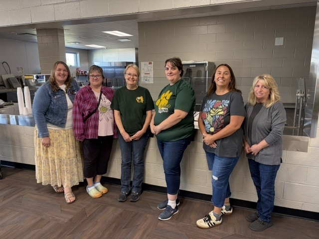 Six women standing side by side in a line inside a school cafeteria, posing for a group photo in front of a service counter.