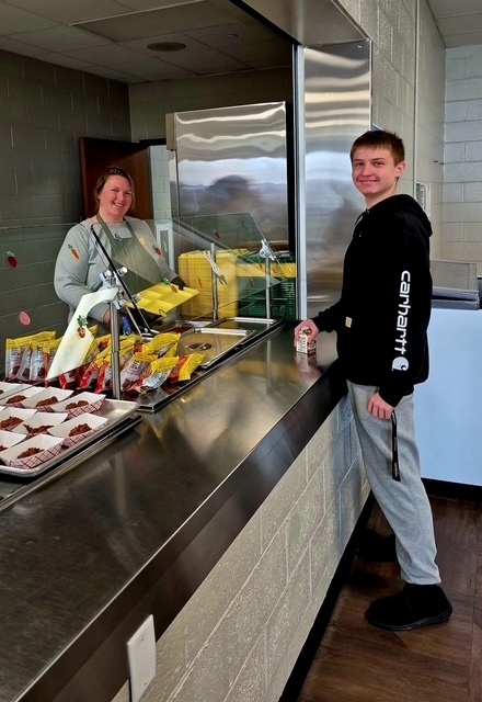 A smiling student stands at a cafeteria counter holding a small carton of milk while a cafeteria worker behind the serving station smiles holding a tray.