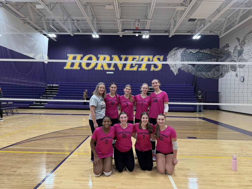 A girls' volleyball team wearing pink "Florence Ladycats" jerseys poses behind a net inside a gymnasium with yellow and purple "HORNETS" branding on the wall.