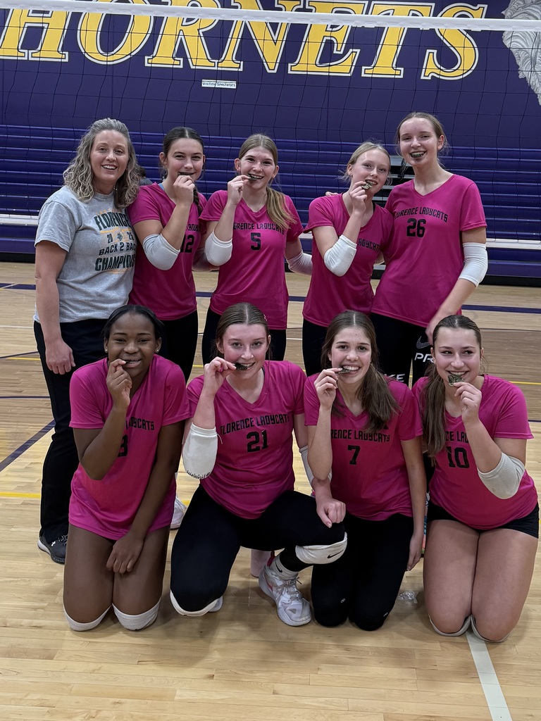 A women's volleyball team wearing pink "Florence Ladycats" jerseys poses on a gymnasium court holding medals in their teeth, with a large "HORNETS" banner behind them.
