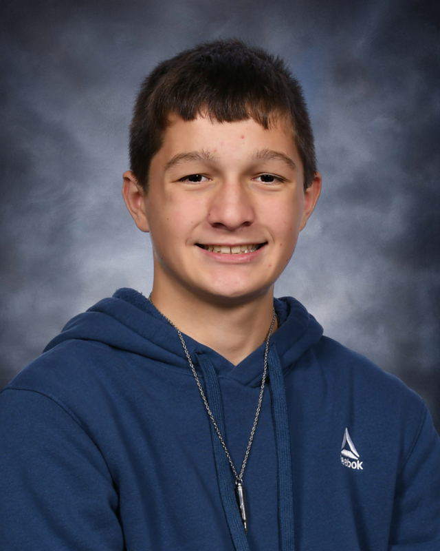 A school portrait of a young boy with short brown hair smiling at the camera. He is wearing a dark blue Reebok hooded sweatshirt and a silver chain necklace with a small pendant. The background is a textured, mottled dark grey and blue canvas.