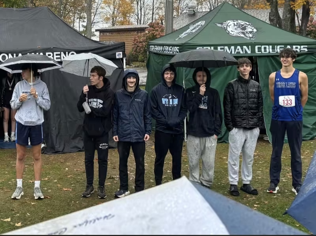 A line of seven young male runners standing outdoors on grass on a rainy day in front of event tents, with several holding umbrellas.
