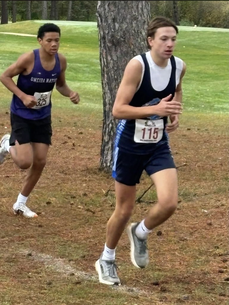 Two young male runners in athletic wear competing in a cross country race along a pine needle-covered grass path.
