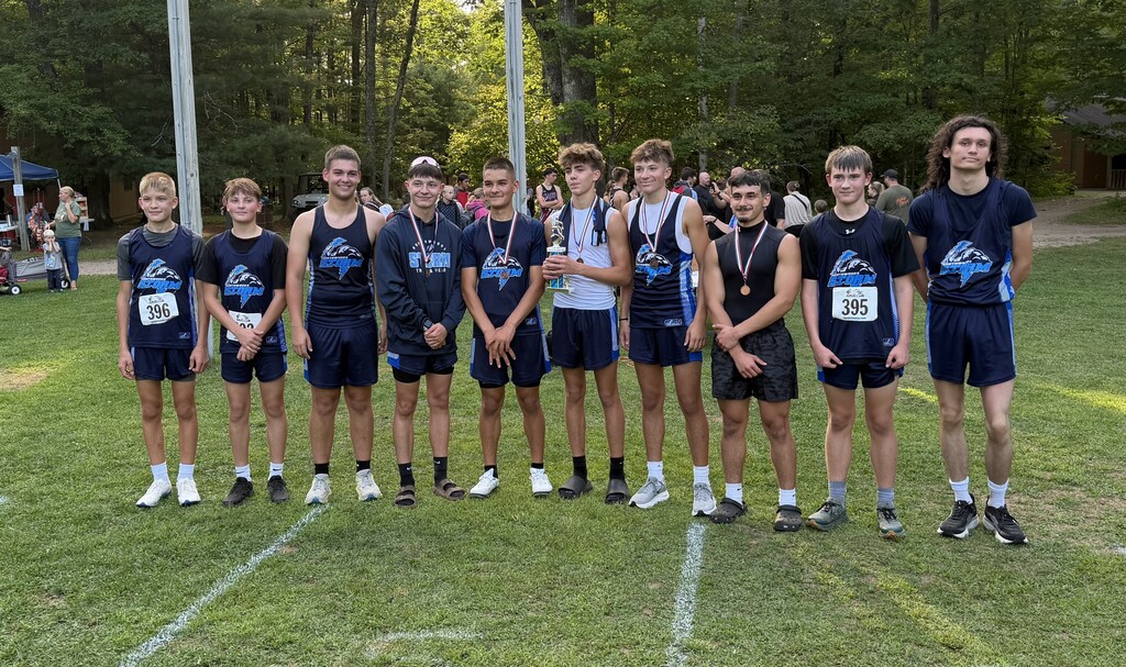 A group of nine male athletes wearing navy blue "Storm" team uniforms standing side-by-side on a grassy field outdoors, with several wearing medals and one holding a trophy.
