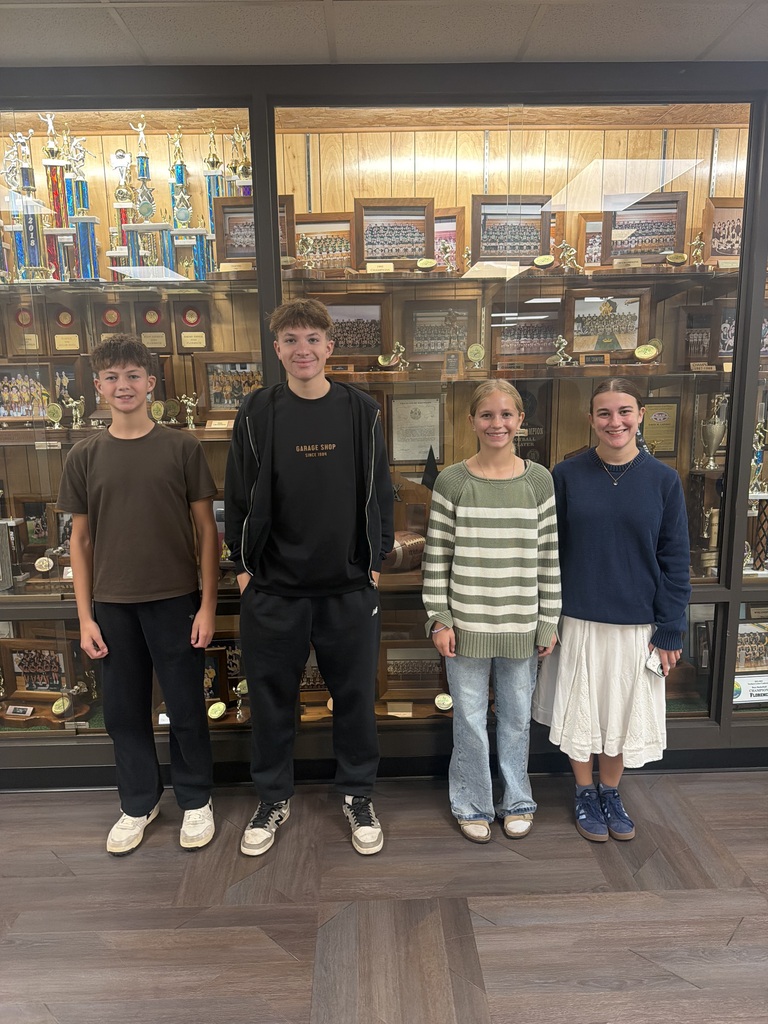 Four young people stand in a line smiling in front of a large glass display case filled with trophies and framed team photos.