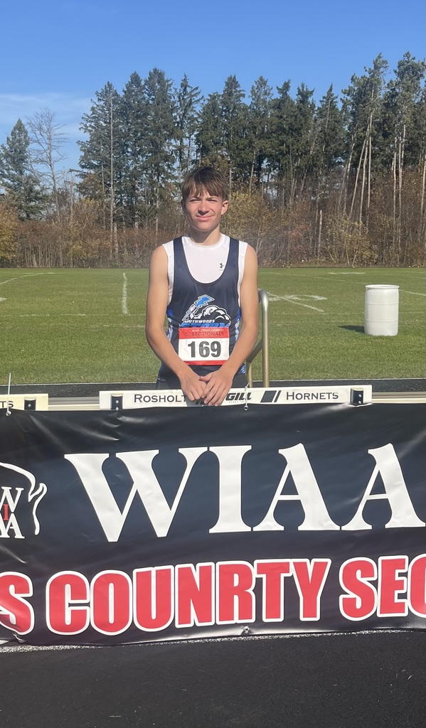 A young male runner wearing a dark blue and white singlet with race bib number 169 poses behind a WIAA Cross Country banner on a bright, sunny day with trees in the background.