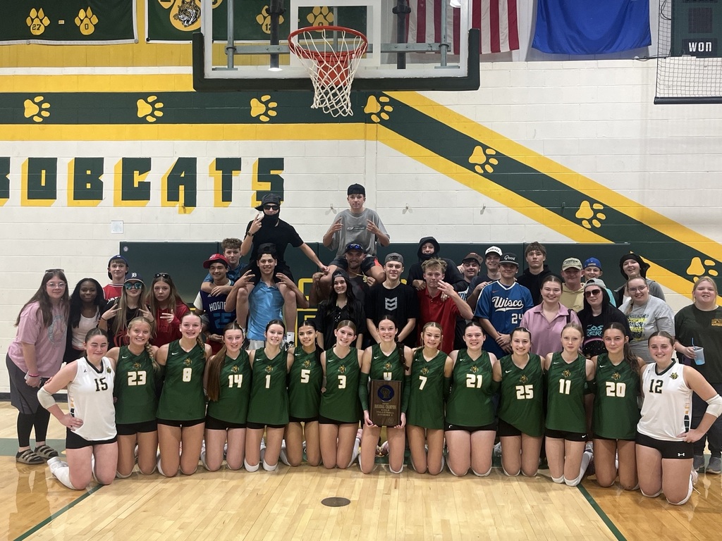 A high school girls' basketball team wearing green and white uniforms kneels in a row on a wooden basketball court, smiling and posing with a championship plaque. Behind them, a group of supportive students, friends, and fans stand in a crowd in front of a gymnasium wall decorated with green and yellow banners and the word "BOBCATS."
