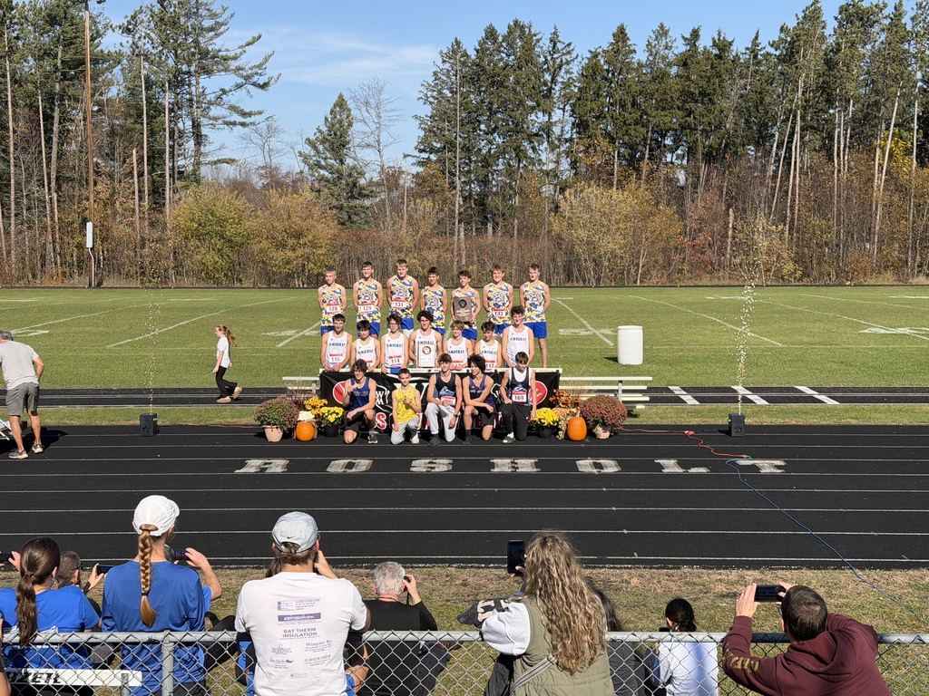 A high school cross-country team poses for a group photograph on a podium during an outdoor award ceremony on a sunny day. The athletes stand in uniform holding trophies, framed by decorative pumpkins and potted flowers on a running track, with spectators in the foreground viewing and taking photos.