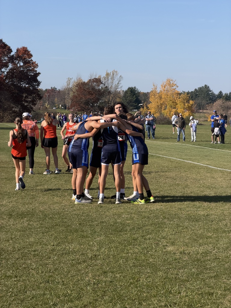 A group of high school cross-country teammates in matching blue uniforms huddle closely together with their arms around each other's shoulders in a circle on a grassy field. Other runners and spectators gather under clear blue skies with autumn trees in the background.
