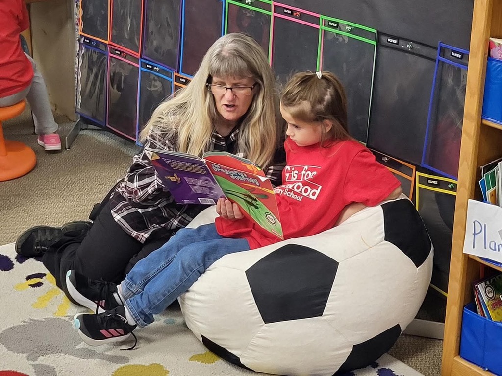 grandma reading to her grandaughter who is sitting on a bean bag chair in a classroom