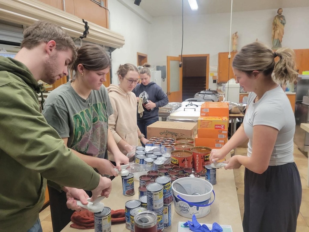students standing around a table, cans of food items, can openers, a bucket, roasters
