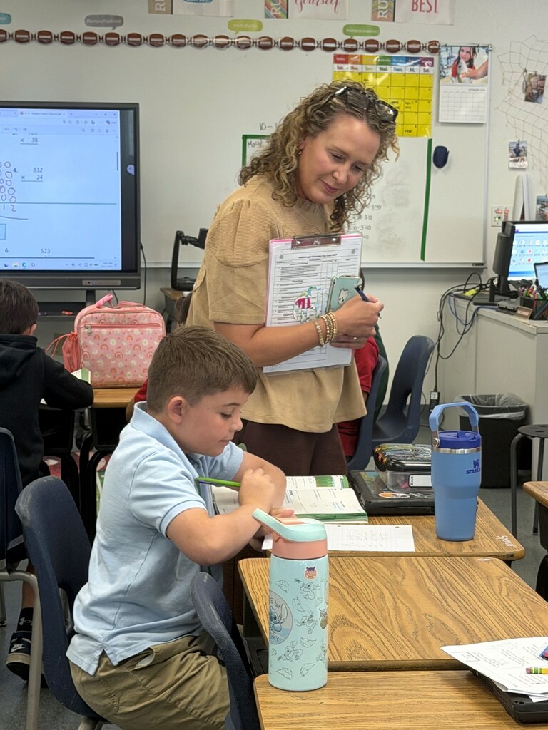 An adult holding a clipboard watches a student work with a pencil in an open workbook during a classroom visit.