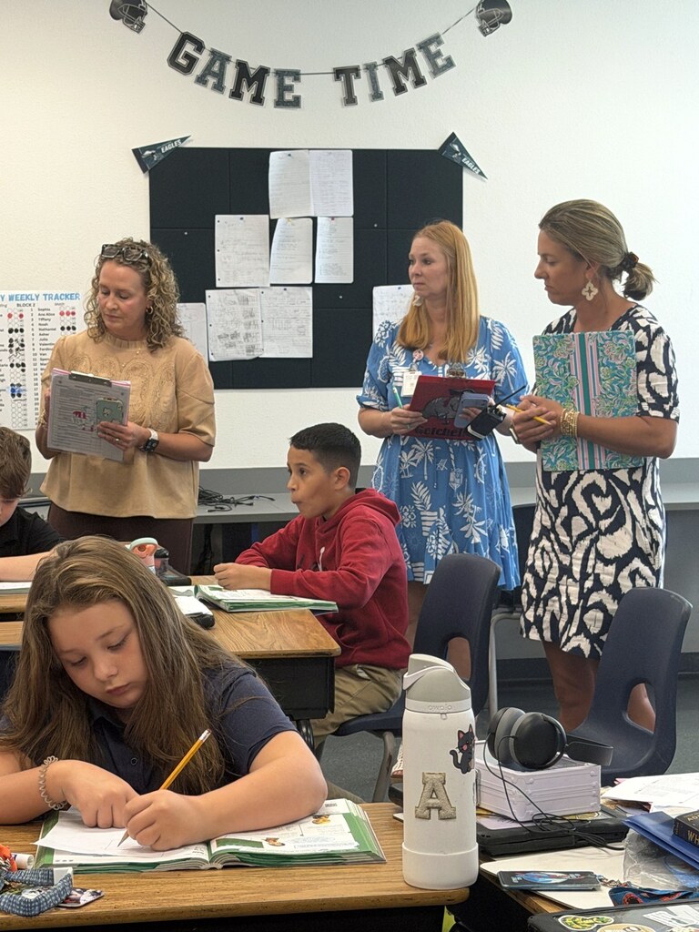 Three adults holding clipboards observe a classroom while students write in open workbooks at their desks.