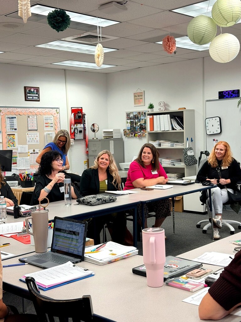 Visiting educators sit around classroom tables with notebooks and papers, smiling and listening during a group discussion.