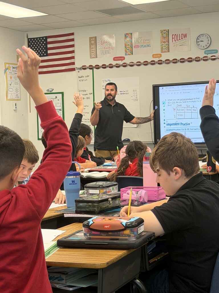 Students raise their hands and work at desks while a teacher leads a math lesson beside a digital display.