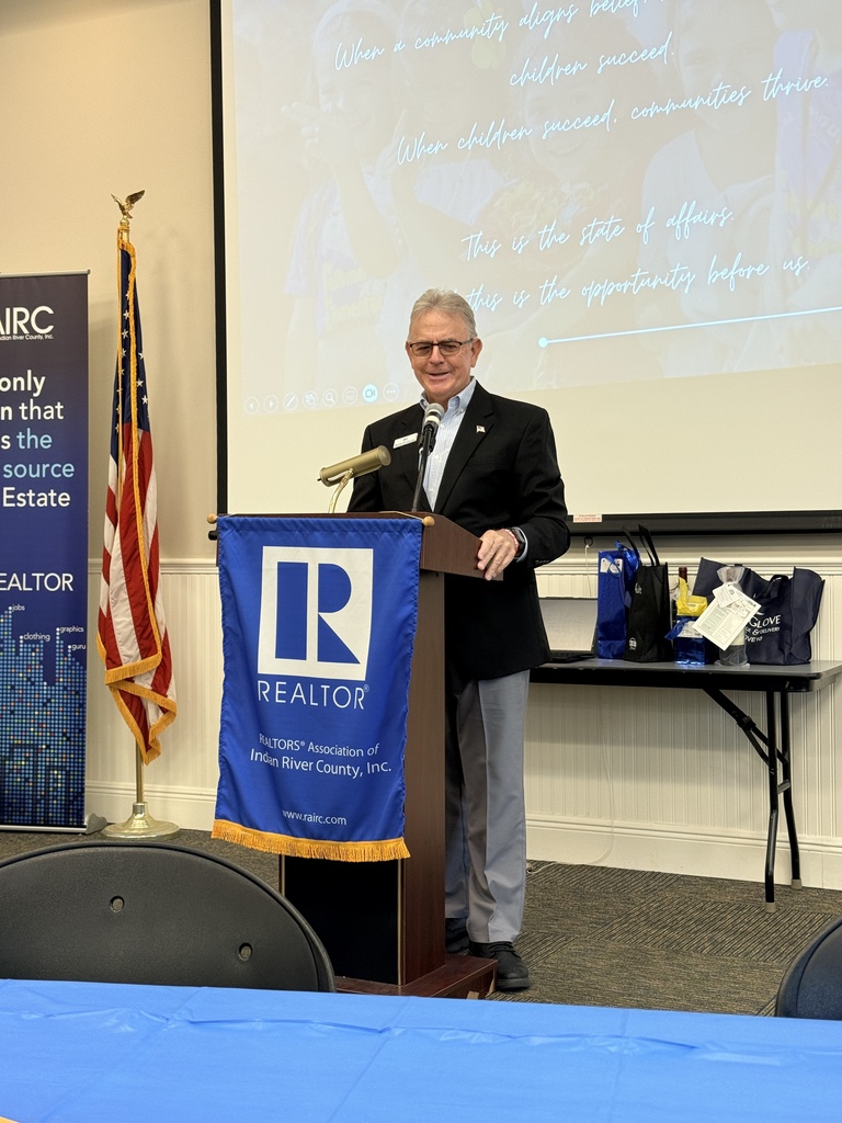 A speaker addresses attendees from a podium displaying the Realtors Association of Indian River County logo. Behind him, a presentation slide emphasizes the connection between community alignment, student success, and thriving communities.