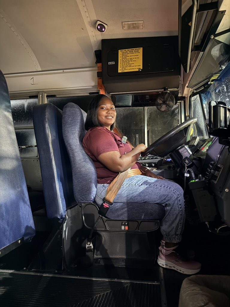 A smiling SDIRC school bus driver sits in the driver's seat of a school bus, wearing her seat belt. The photo shows the driver's area, steering wheel, dashboard, and rows of blue passenger seats behind her.