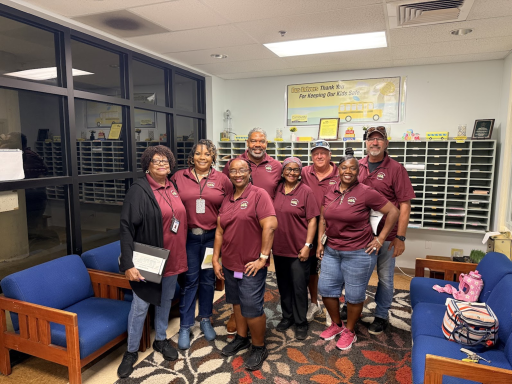 Eight SDIRC transportation team members pose together for a group photo inside a transportation office. Most are wearing matching maroon district polo shirts. A banner behind the group reads, “Bus Drivers Thank You For Keeping Our Kids Safe.”