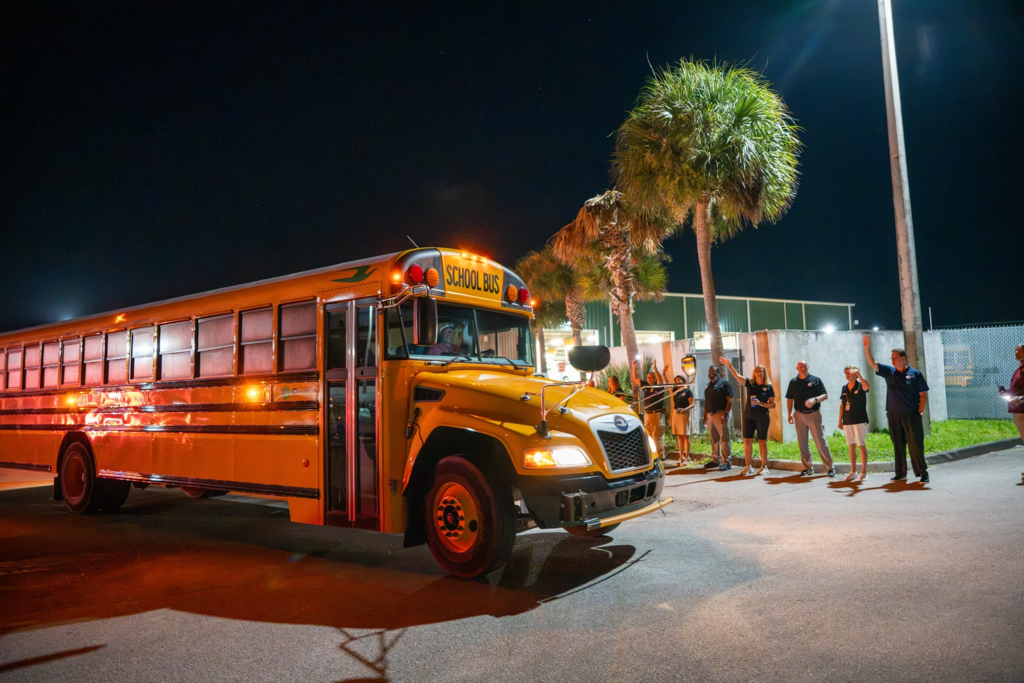 A yellow SDIRC school bus travels along a roadway at night with its headlights and exterior lights illuminated. A group of people stands along the roadside, smiling and waving as the bus passes. Palm trees and outdoor lighting are visible in the background.