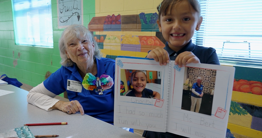 A volunteer smiles next to a young girl holding up a book.