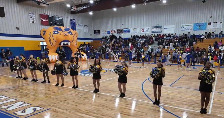 Cheerleaders smiling at a pep rally in the school gymnasium.