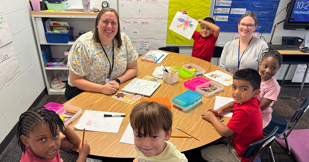 A group of smiling students sitting around a small table with two teachers.