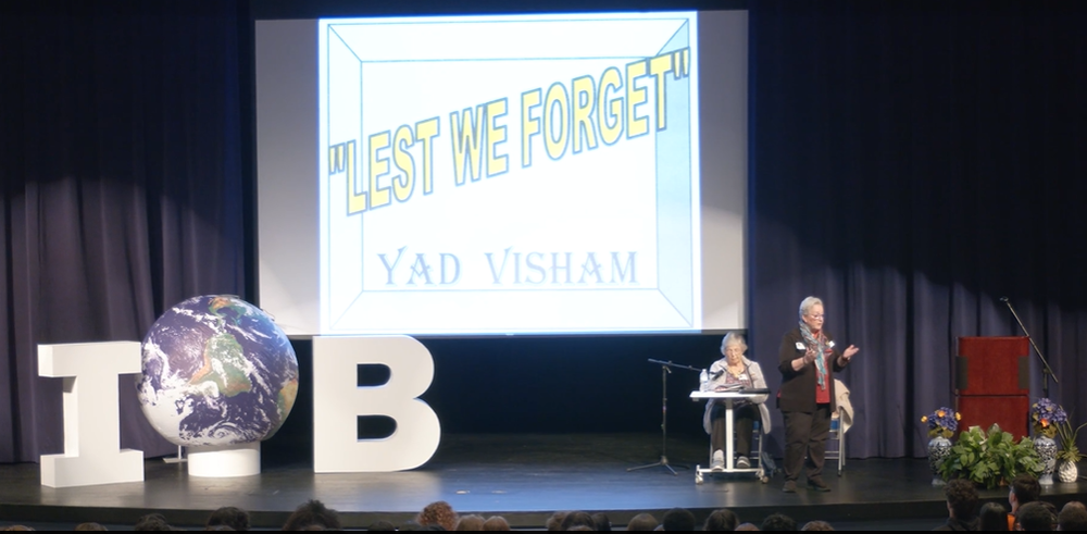 A special guest speaking on a high school stage with the words "Lest We Forget" behind her.