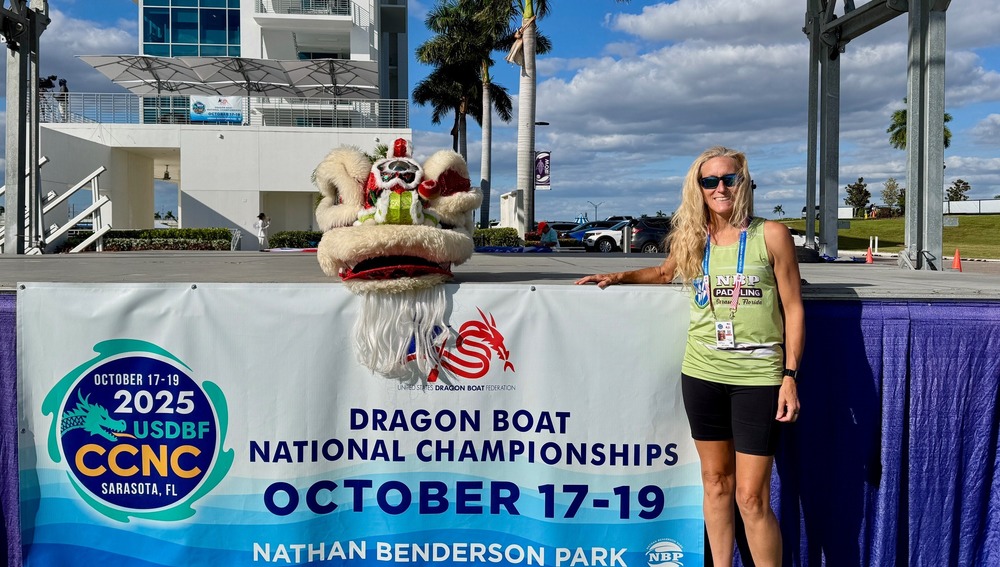 A woman in green shirt and black shorts standing next to a banner that reads, Dragon Boat National Championships October 17-19 at Nathan Benderson Park