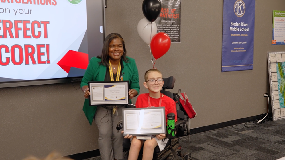 A school principal stands next to a student in a wheelchair, celebrating his two perfect scores on state assessments.