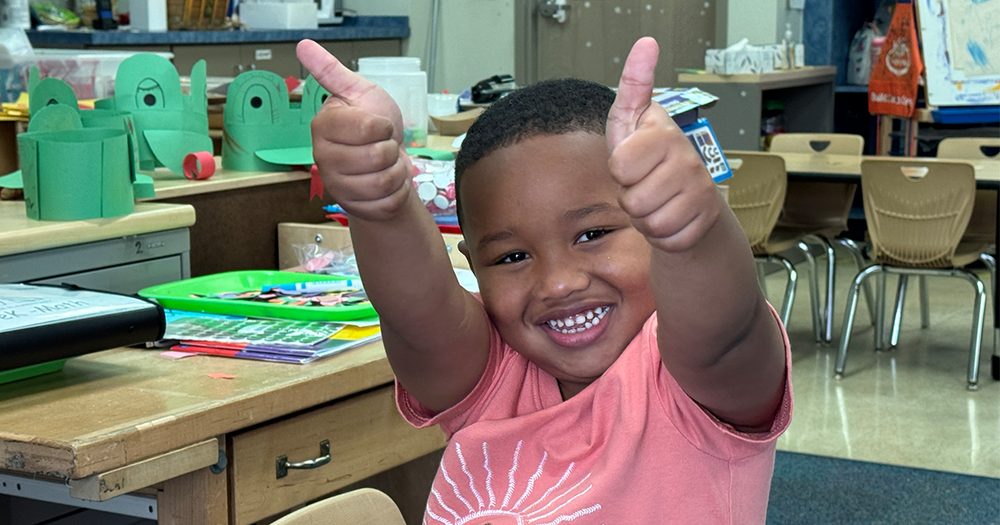  A smiling boy in a pink shirt gives two thumbs up in a classroom.