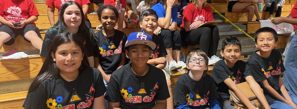 students wearing black tshirts that read "STEM Club" and smiling while sitting on wooden bleachers.