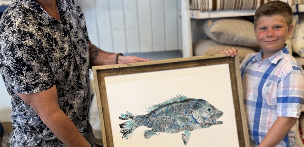 A young boy wearing a plaid shirt holds a wooden frame with fish print.