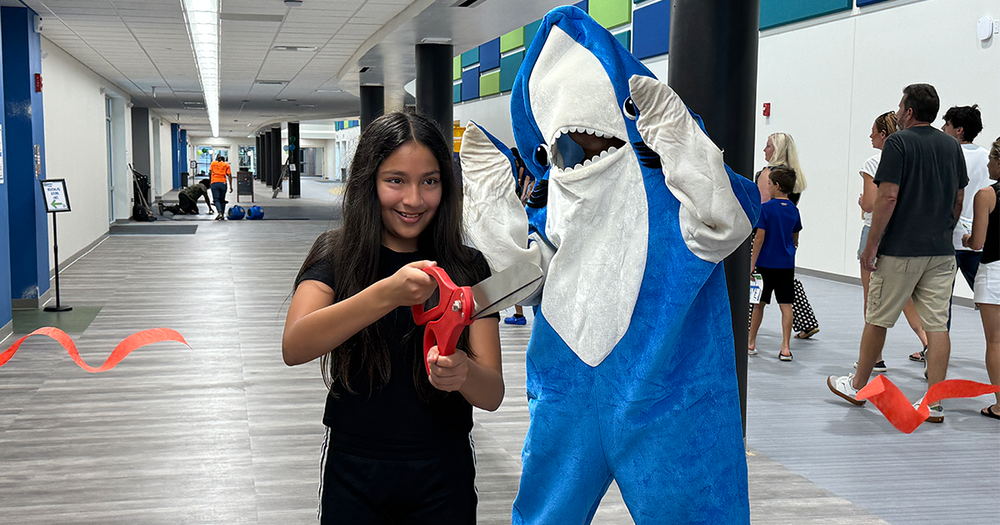 A girl cuts a red ribbon with large scissors next to a shark mascot during a school grand opening event.