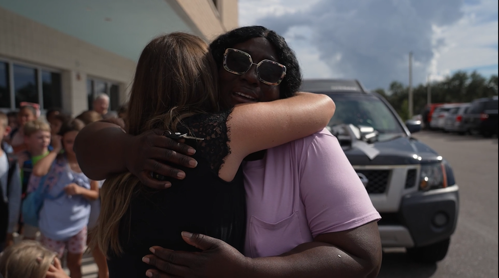 Two women hugging in front of SUV with a large silver bow on its hood.