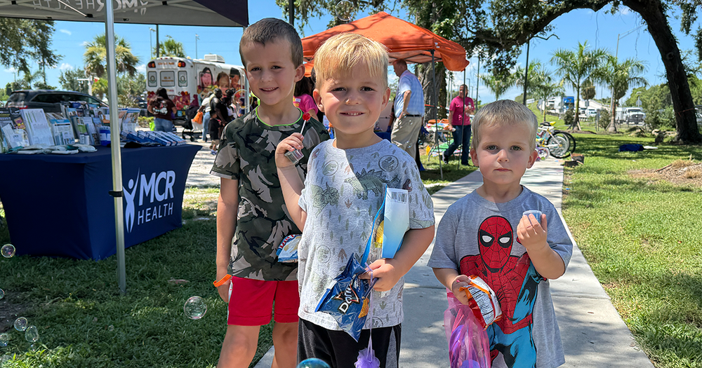 Three little boys smiling while holding bags and free items distributed during a community event.