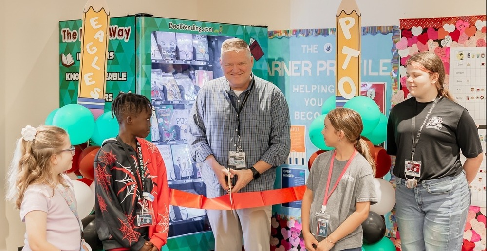 Dr. Grimes is cutting the ribbon  to officially open our new book vending machine with 4 students looking on.